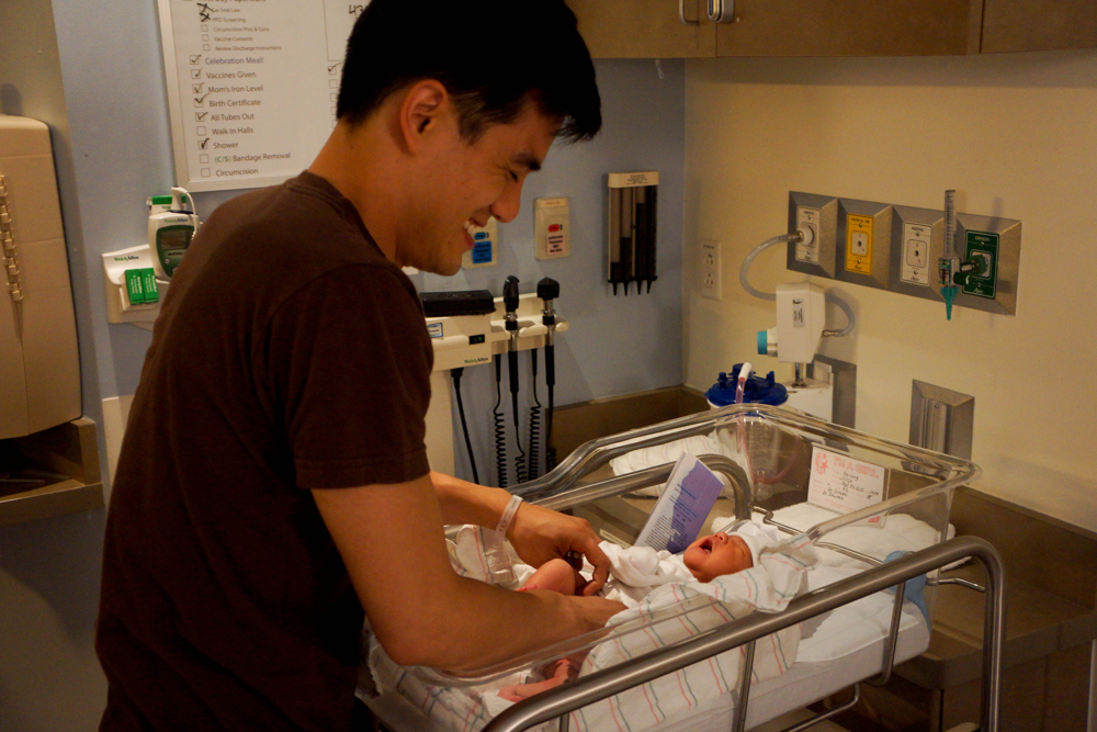 A new father smiles while changing his newborn baby’s diaper in the hospital, illustrating how partners can actively share baby care duties.